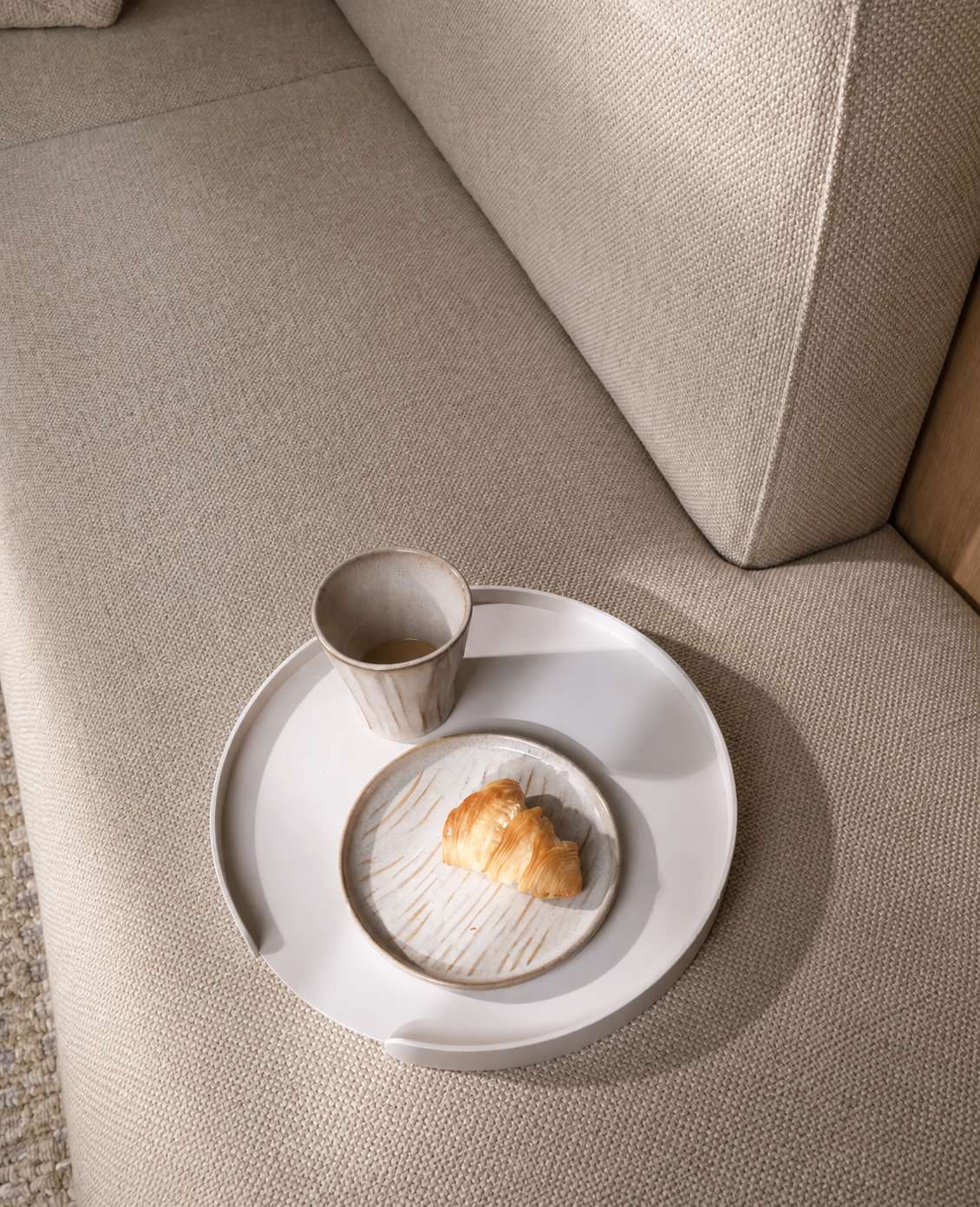 Weißes rundes Metalltablett Milo von Siqual Design auf hellem Stoffsofa, fotografiert aus der Draufsicht mit Kaffeebecher, Keramikteller und Croissant in moderner Wohnatmosphäre.
