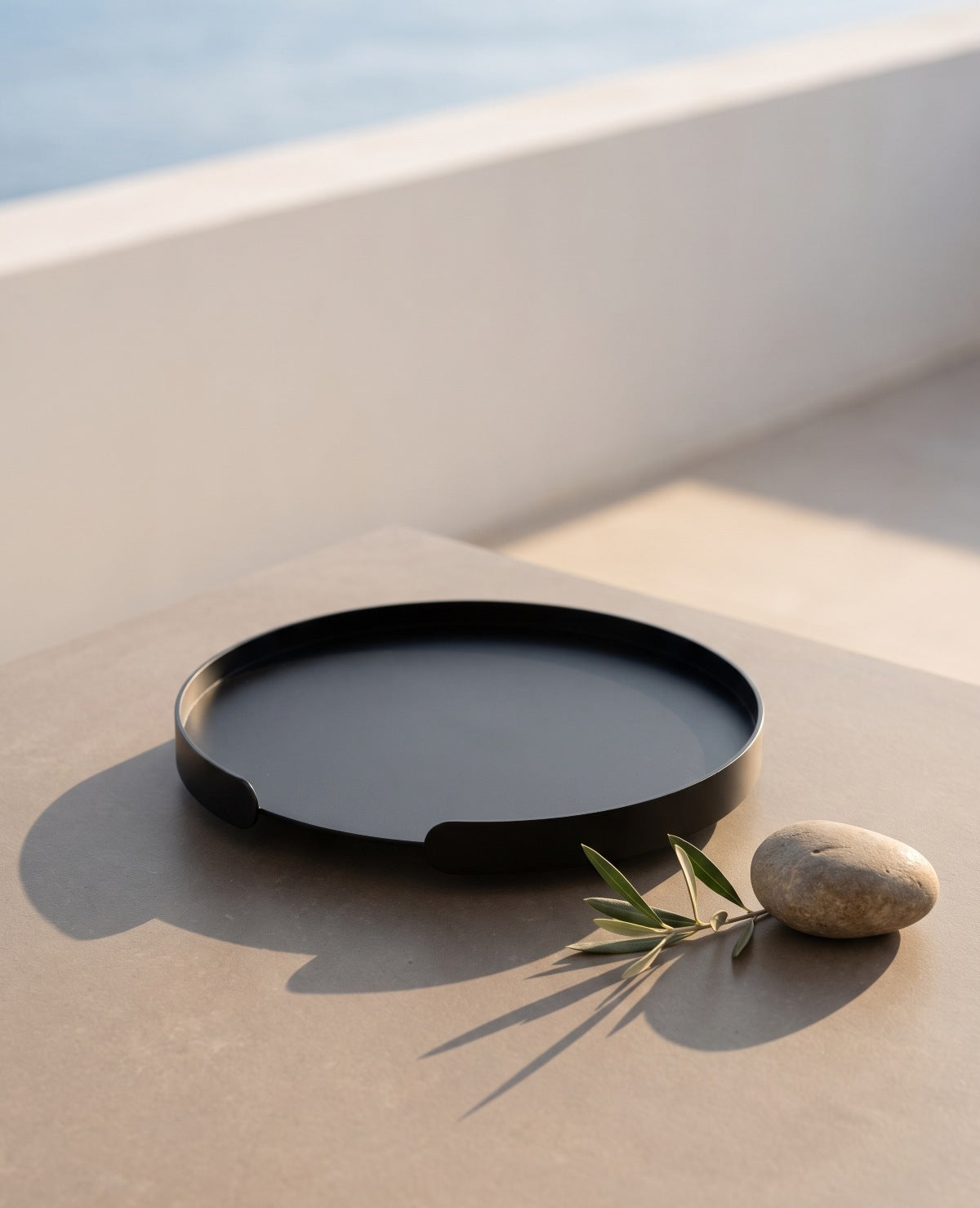 Schwarzes rundes Metalltablett Milo von Siqual Design auf hellem Steintisch mit Olivenzweig, Keramikbecher und mediterraner Dekoration.