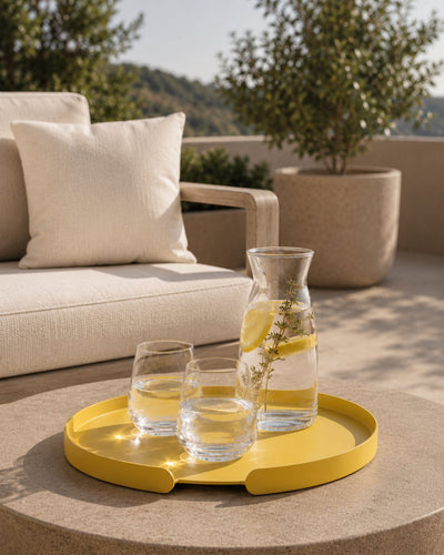 Gelbes rundes LILO Tablett von Siqual Design auf einem Outdoor-Tisch mit Wassergläsern, Zitronen-Karaffe und moderner Lounge im Hintergrund.